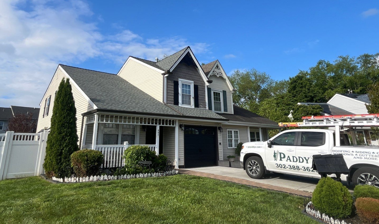 Two story home with gray siding and white trim, lawn and a Paddy's Roofing work truck on the driveway.